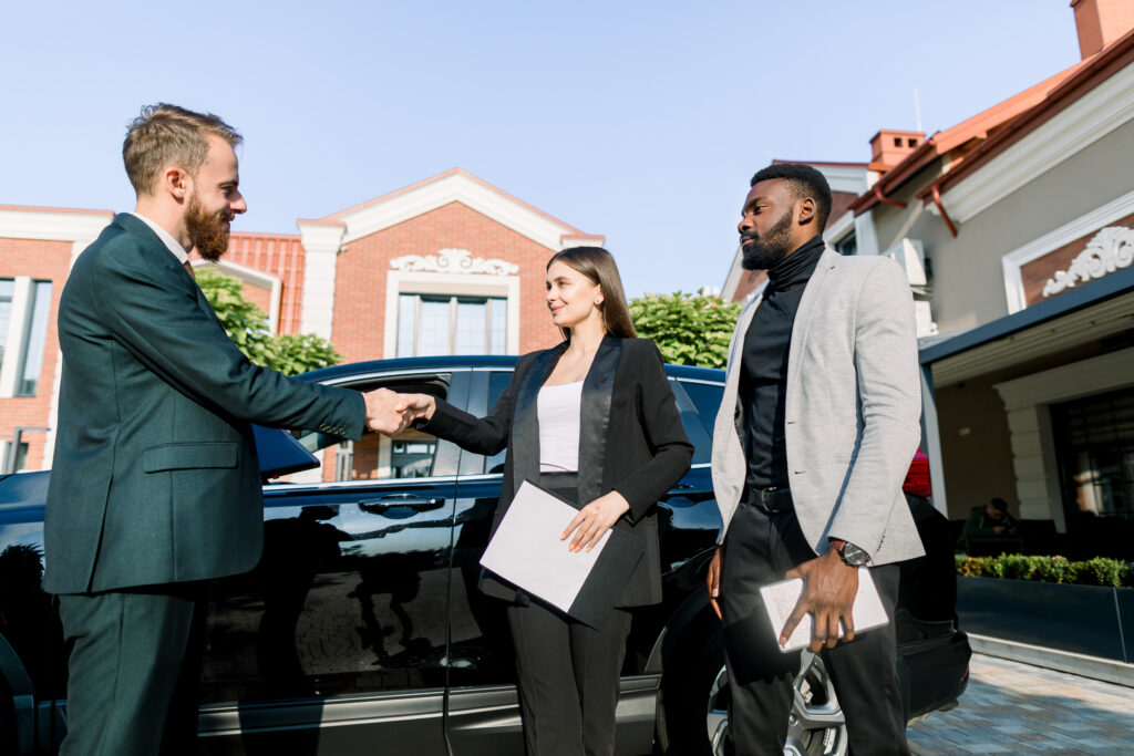 Welcome to our team. three young multiethnical business people standing near car outdoors. happy young woman shaking hands with new colleague man, african man holding tablet stands nearby